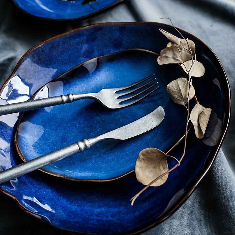 Atlas Dinner Plate: Deep blue ceramic dinner plate with organic wavy rim, stacked with a smaller plate, fork, and knife.