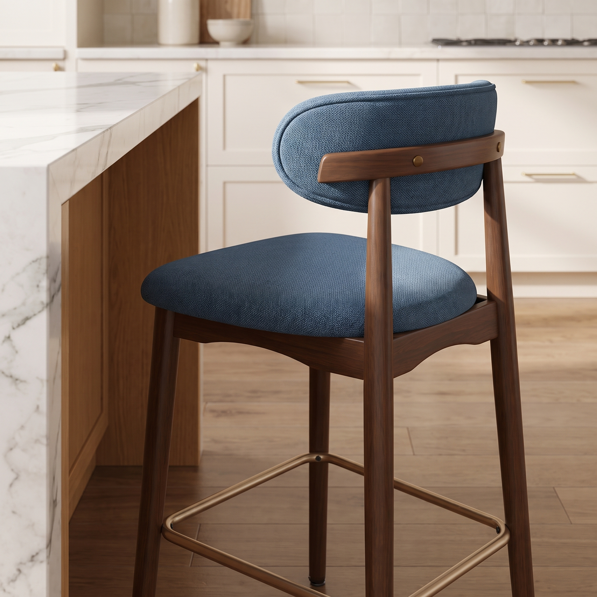 A modern bar stool with a blue upholstered seat and backrest, a medium brown wooden frame, and a gold metal footrest, set in a bright kitchen next to a marble island.