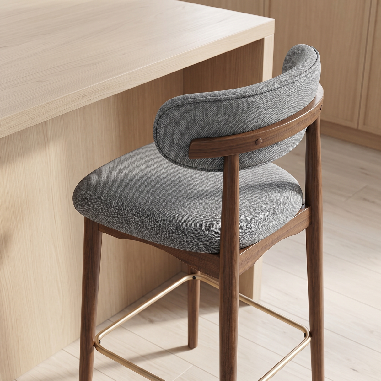 A modern bar stool with a gray upholstered seat and curved backrest, supported by a medium brown wooden frame with a gold metal footrest, shown next to a light wood counter in a kitchen setting.