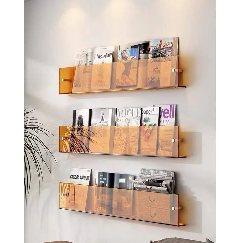 Three amber acrylic wall-mounted magazine shelves holding various magazines on a white wall.