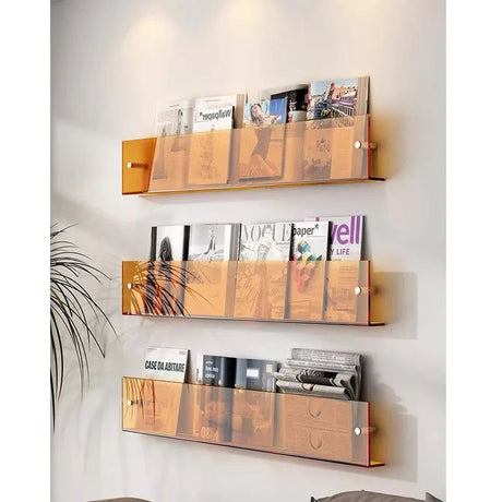 Three amber acrylic wall-mounted magazine shelves holding various magazines on a white wall.