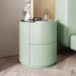 Mira Bedside Table in light green with two curved drawers and a round top, next to a bed.