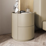 Mira Bedside Table in khaki, a round nightstand with two curved drawers, next to a bed.
