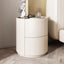 Mira Bedside Table in off-white, round with two drawers, next to a bed on a wooden floor.