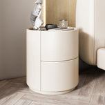 Mira Bedside Table in off-white, round with two drawers, next to a bed on a wooden floor.
