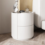 White Mira bedside table with two drawers and a round base, next to a bed.