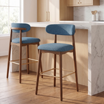 Two upholstered bar stools with blue fabric seats and backs, wooden legs, and gold-tone footrests, shown in a bright kitchen setting with a marble island and wood floor.
