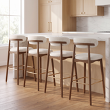 Four modern bar stools with white upholstered seats and backs, dark wood legs, and gold-tone footrests, arranged around a kitchen island in a bright, contemporary kitchen.