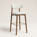 A modern bar stool with a light beige upholstered seat and curved backrest, supported by a medium brown wooden frame with a gold metal footrest, set against a solid light beige background.