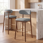 Two upholstered bar stools with gray fabric seats and backs, dark wood legs, and gold-tone footrests, shown in a bright kitchen setting beside a marble island.