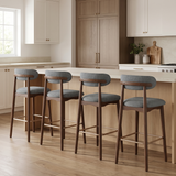 Four modern bar stools with gray upholstered seats and backs, dark wood legs and armrests, and brass footrests, arranged around a light wood kitchen island in a bright, contemporary kitchen.