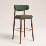 A modern bar stool with a green upholstered seat and curved backrest, supported by a medium brown wooden frame with a gold metal footrest, set against a plain light beige background.