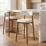 Two modern bar stools with cream upholstered seats and backs, dark wood frames, and gold-tone footrests, positioned in front of a white marble kitchen island in a bright kitchen setting.
