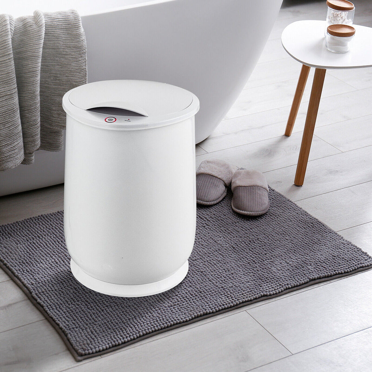 White towel warming bucket on a gray bath mat with slippers and a white bathtub in the background.