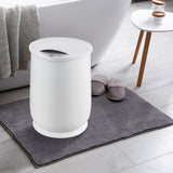 White towel warming bucket on a gray bath mat with slippers and a white bathtub in the background.